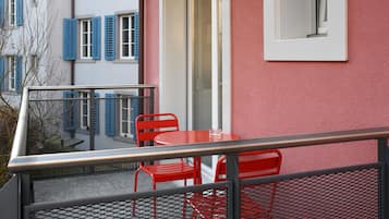 Un balcón con sillas de metal rojas y una mesa, con vista a un edificio con contraventanas azules y una pared rosa.