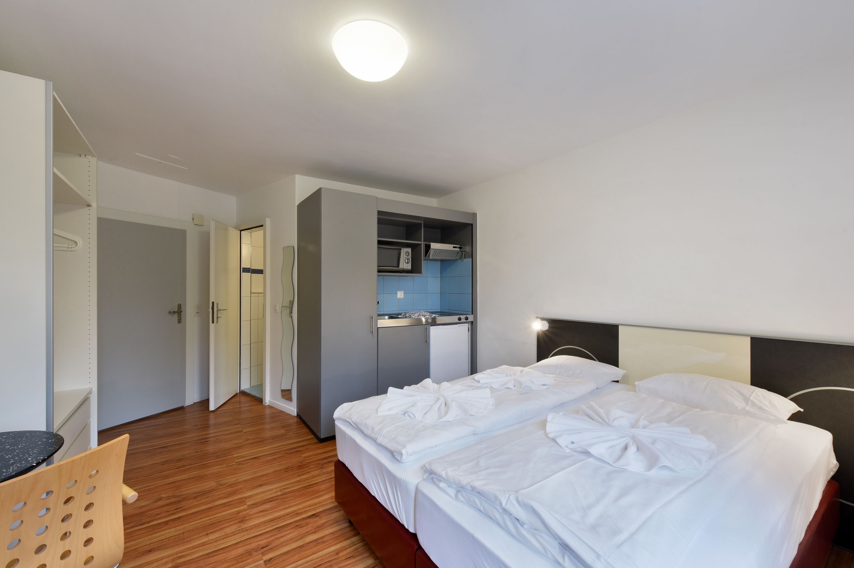A hotel room with a bed, a desk, a chair, a kitchenette, and a bathroom.