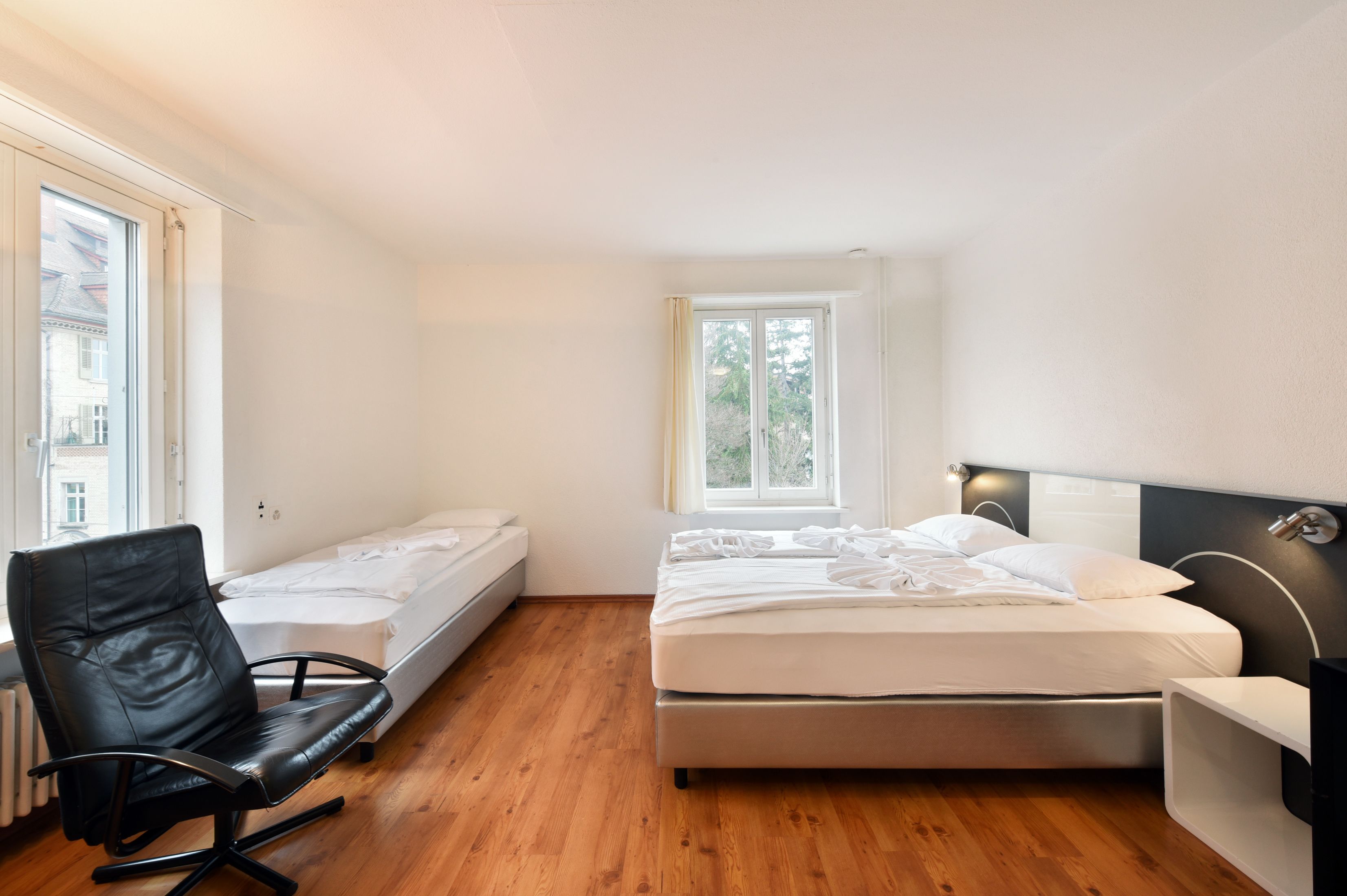 A small, modern room with a wooden floor, a black leather chair, a bed, and a window with a view of buildings outside.