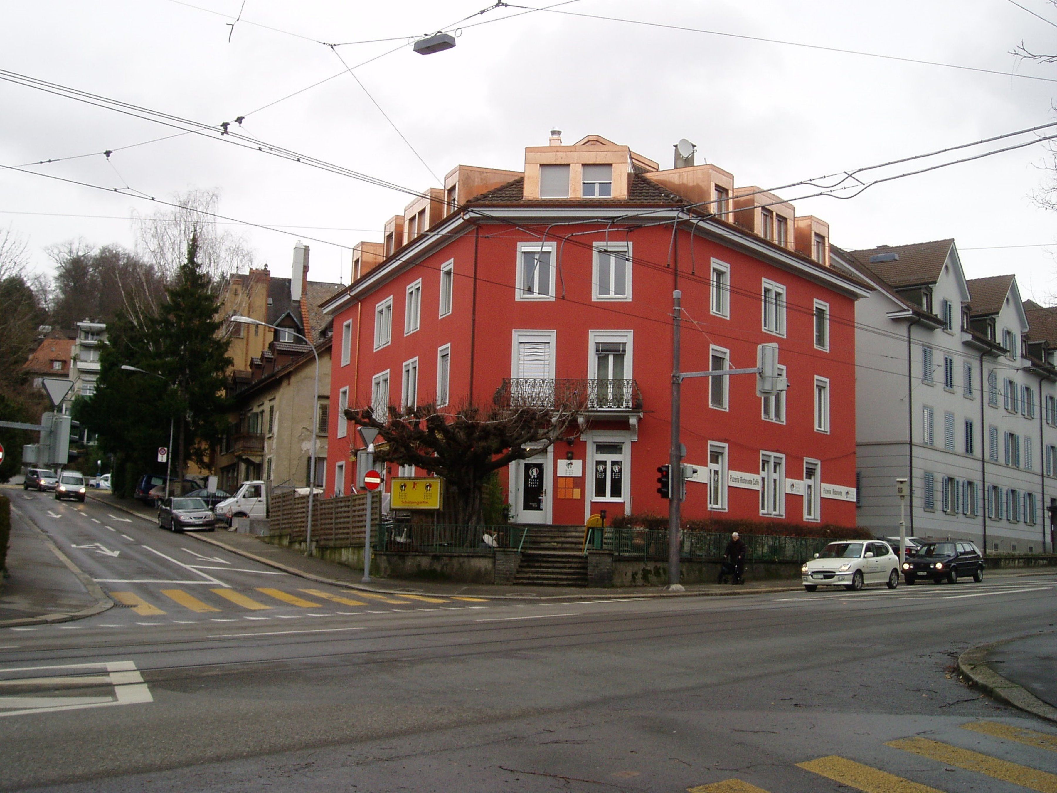 Un edificio rojo con múltiples ventanas y un balcón, situado en una esquina de calle con paso de peatones.