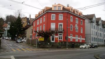 Un edificio rojo con múltiples ventanas y un balcón, situado en una esquina de calle con paso de peatones.