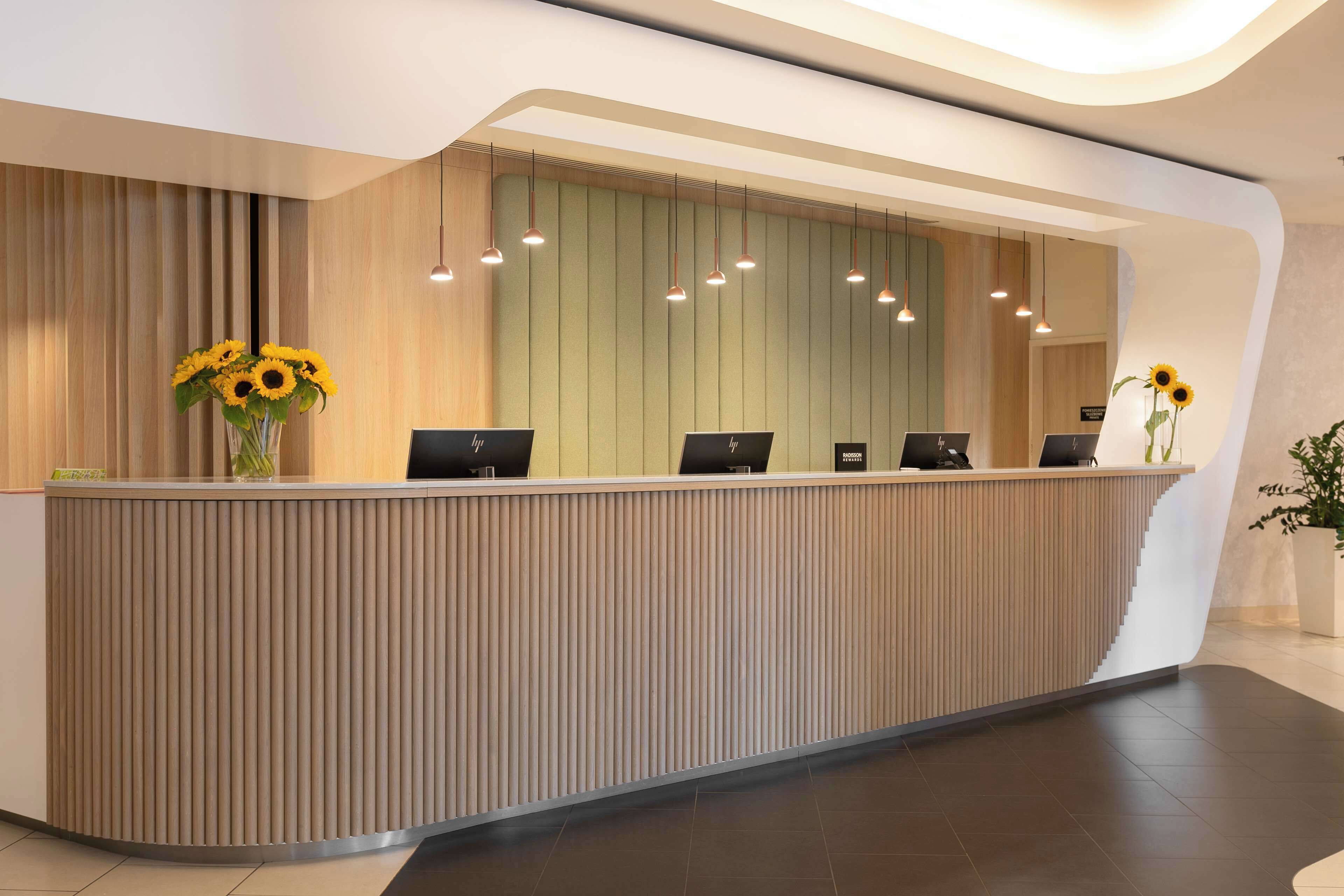 Un mostrador de recepción de diseño moderno, con un panel de madera de fondo, varias estaciones de trabajo con computadoras y un jarrón con girasoles.