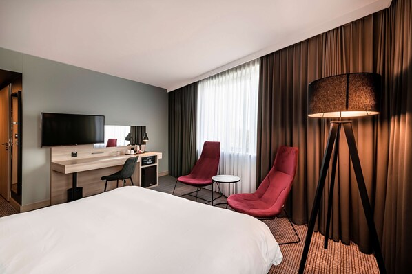 Habitación de hotel con una cama grande, un escritorio con televisor de pantalla plana, dos sillas rojas, una mesita redonda y una lámpara de pie.