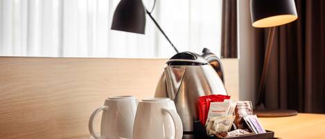 Una bandeja con dos tazas blancas, una tetera eléctrica plateada y una bandeja de bocadillos sobre una mesa de madera en una habitación de hotel.
