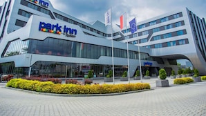A modern building with the 'Park Inn' sign, featuring a glass facade, multiple flags, and landscaped areas with shrubs. -  Park Inn by Radisson Krakow (Kraków)