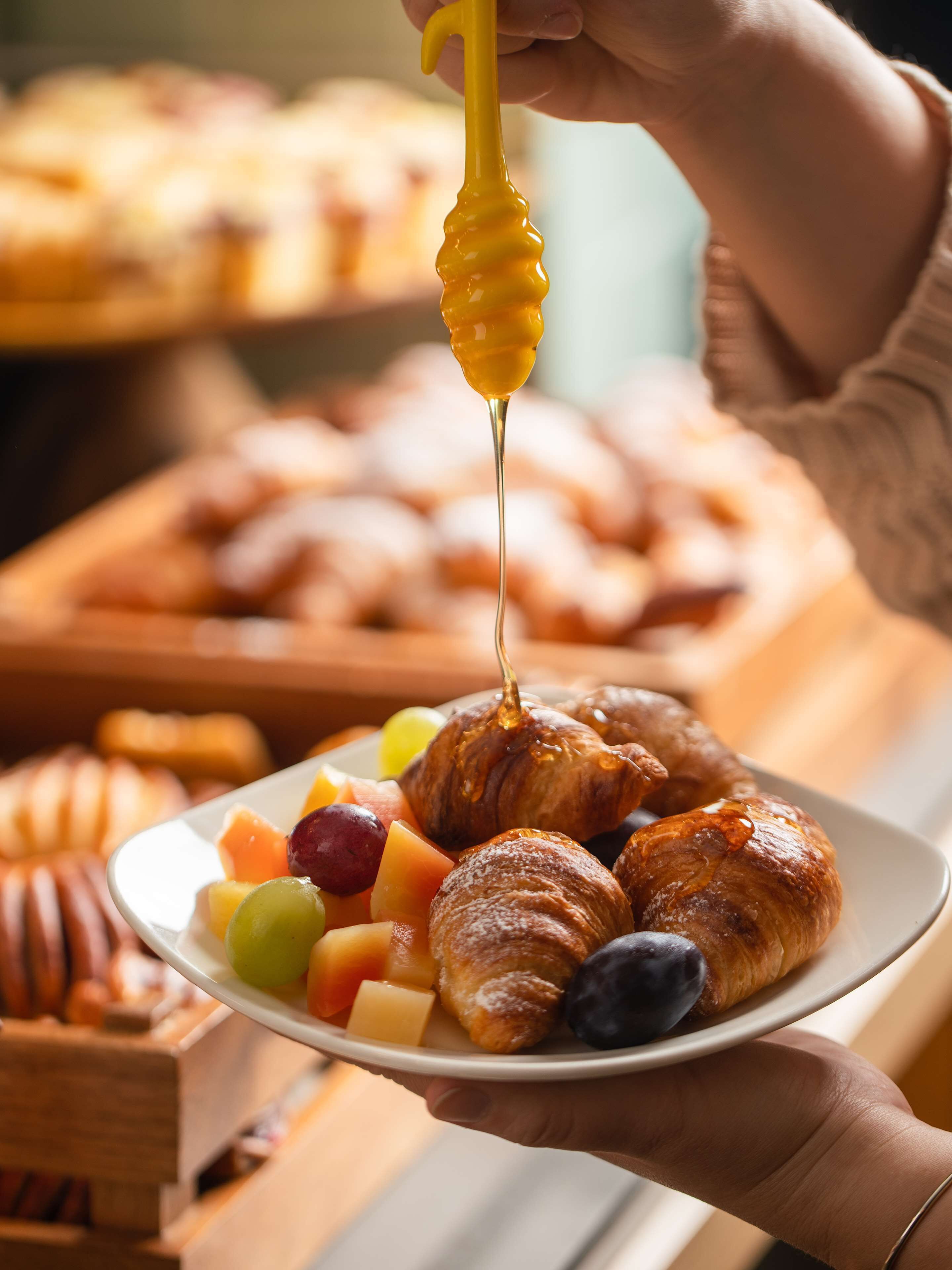 Ein Teller mit Croissants und Früchten, und eine Hand, die einen Honiglöffel hält.