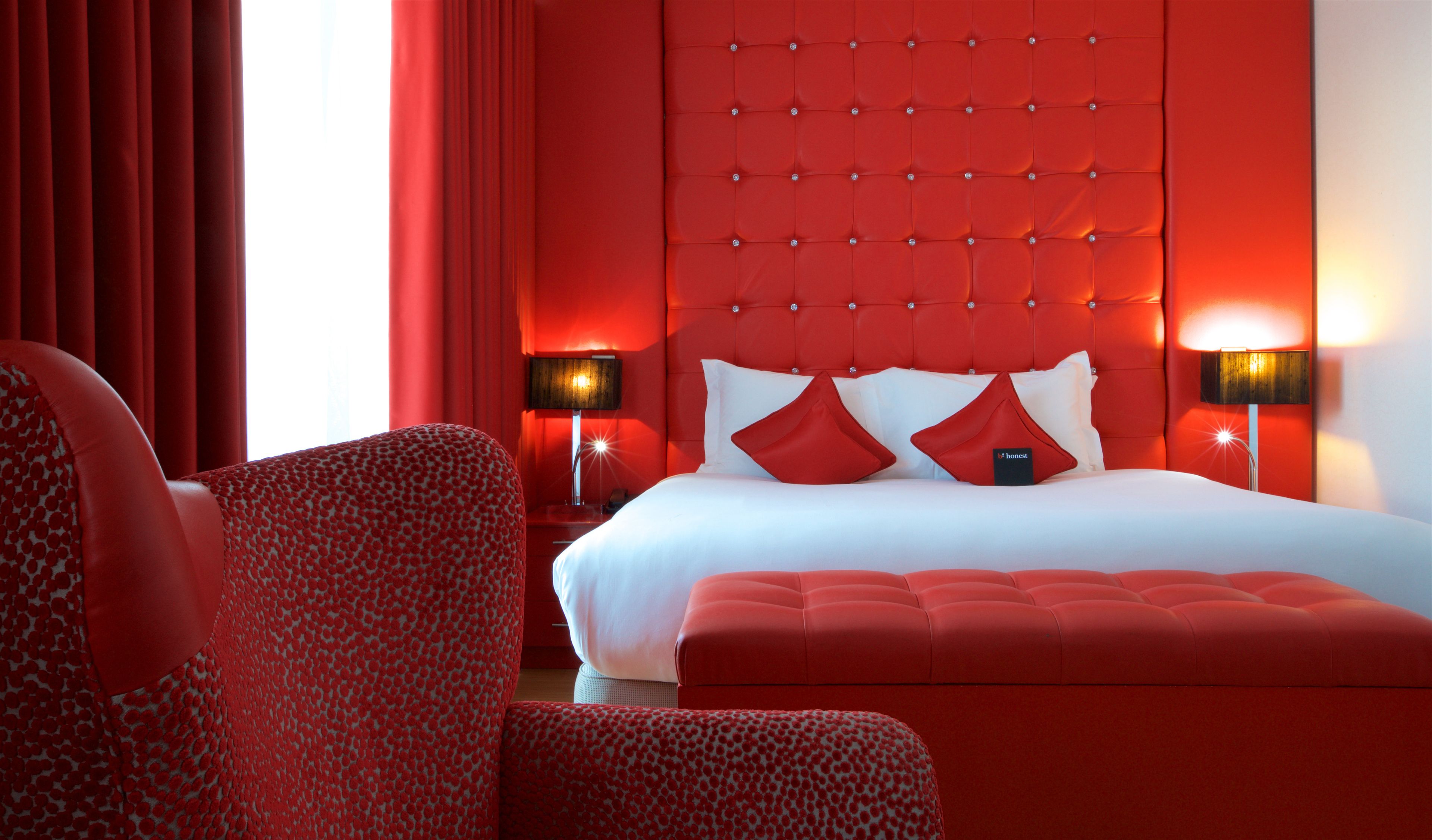 A hotel room with a red-themed decor, including a tufted headboard, red curtains, and matching red furniture.