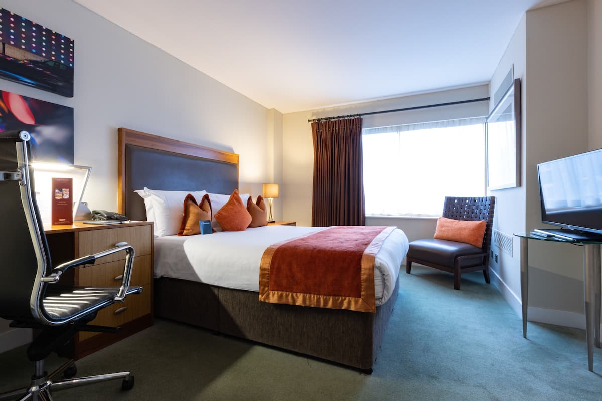 Une chambre d’hôtel comprenant un lit, un bureau avec une chaise, une télévision et une fenêtre avec des rideaux.