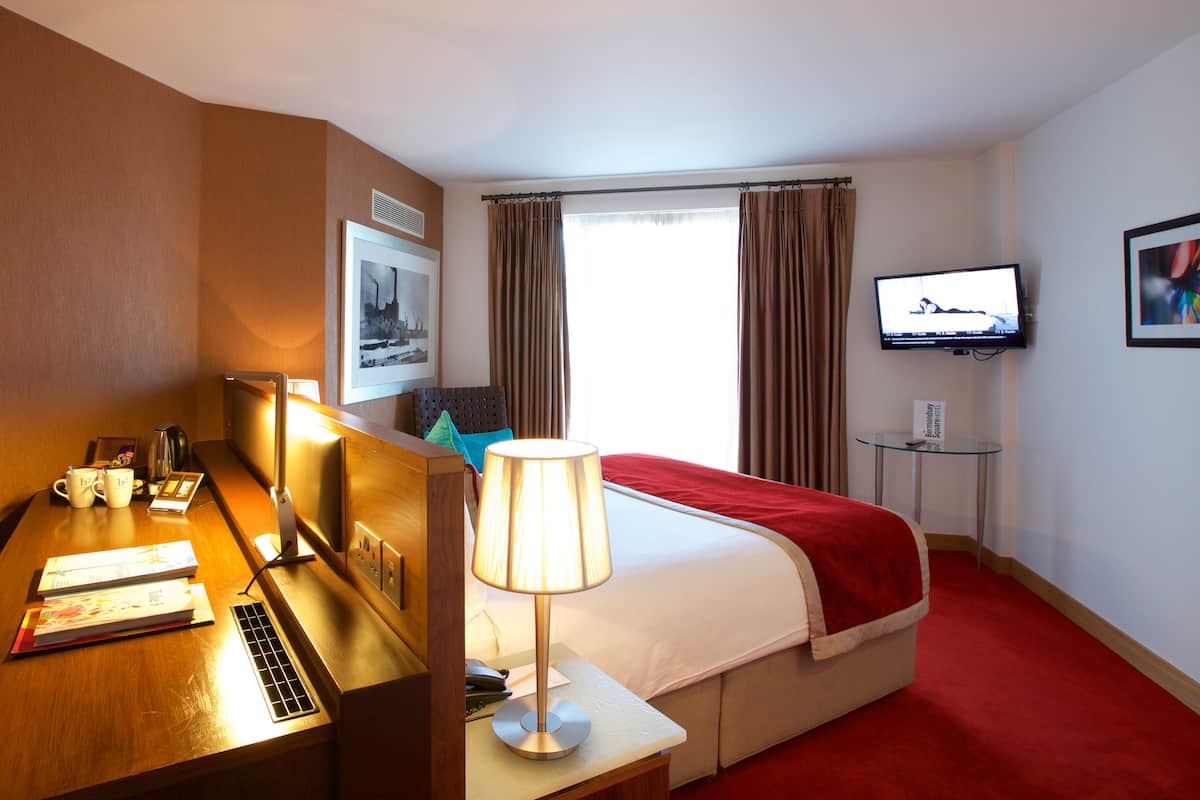 Une chambre d’hôtel avec un lit, un bureau avec une lampe, une télévision et une fenêtre avec des rideaux.