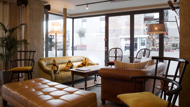 Ein modernes Wohnzimmer mit Sofa, Sesseln und Couchtisch. Große Fenster bieten Blick auf die Straße.