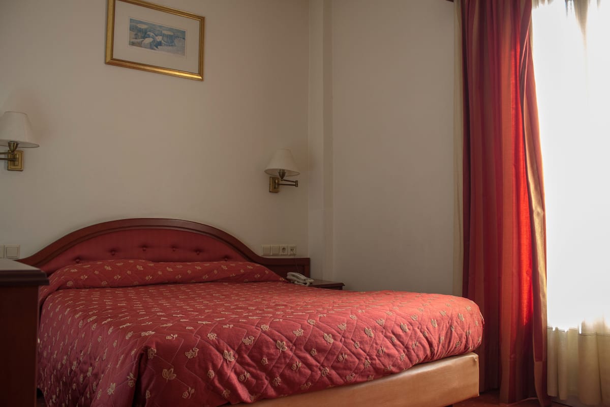 A neatly made bed with a red quilt, two bedside lamps, a framed picture on the wall, and a window with red curtains.