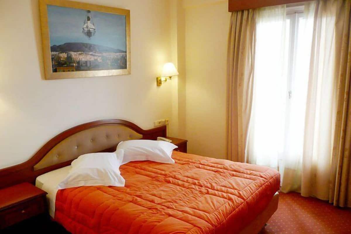 A hotel room with a bed, a nightstand, a lamp, a painting on the wall, and a window with curtains.