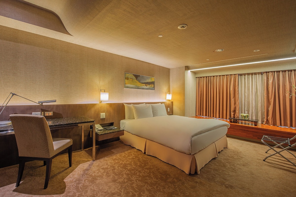 Quarto de hotel com cama, escrivaninha, cadeira e uma grande janela com cortinas.