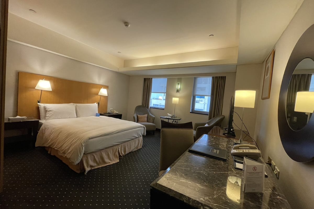 Quarto de hotel com uma cama grande, duas mesas de cabeceira, uma escrivaninha com cadeira, abajur, espelho e janela com cortinas.