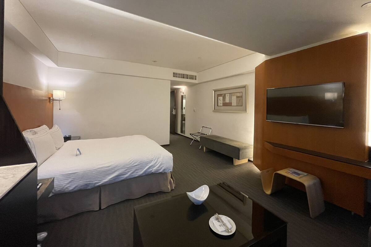 Quarto de hotel com uma cama grande, uma escrivaninha, uma cadeira, uma televisão e uma mesinha com uma tigela e um prato.