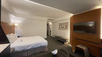 Quarto de hotel com cama, escrivaninha, cadeira, televisão e uma pequena mesa com uma tigela e um prato.