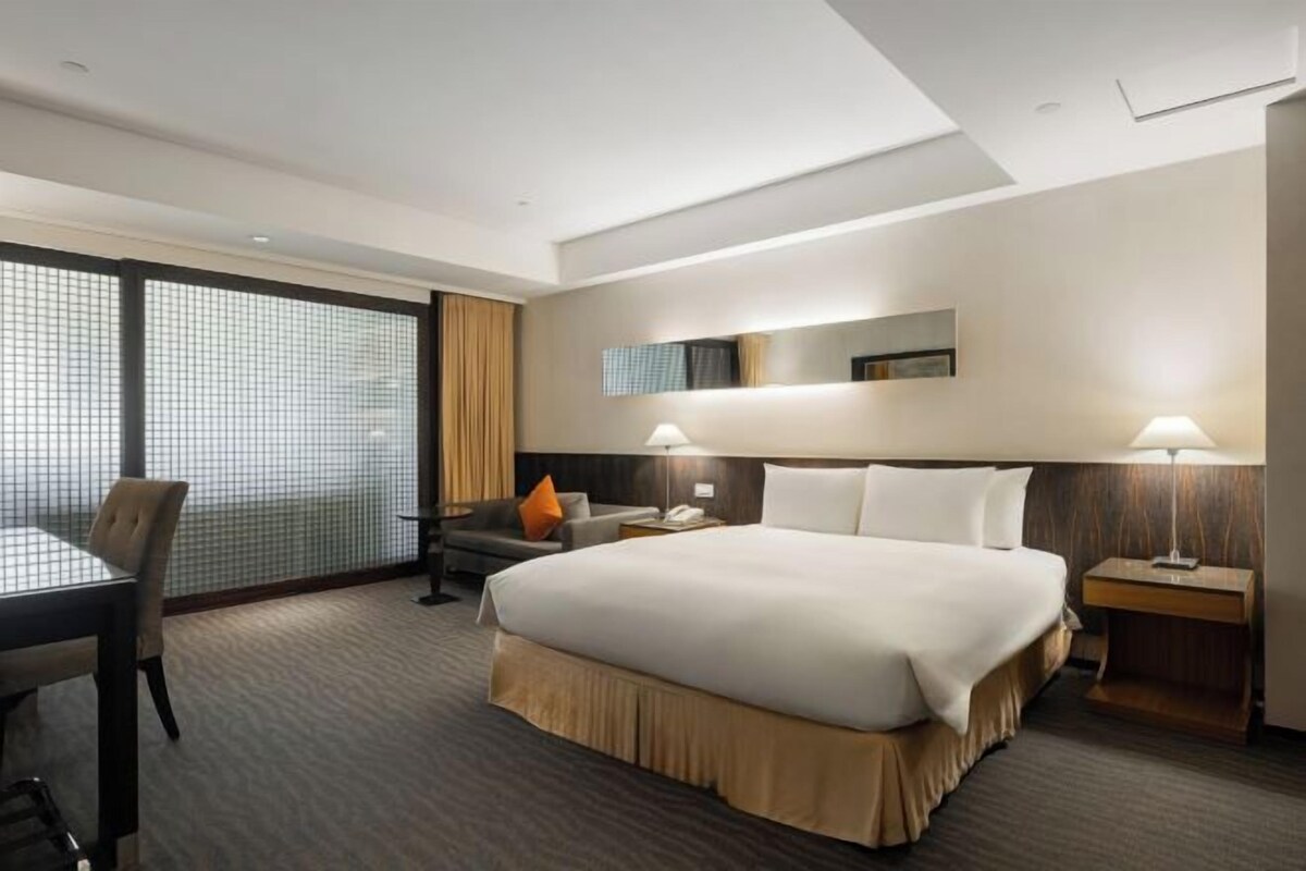 A hotel room with a large bed, a desk, two lamps, a chair, and a TV on the wall.