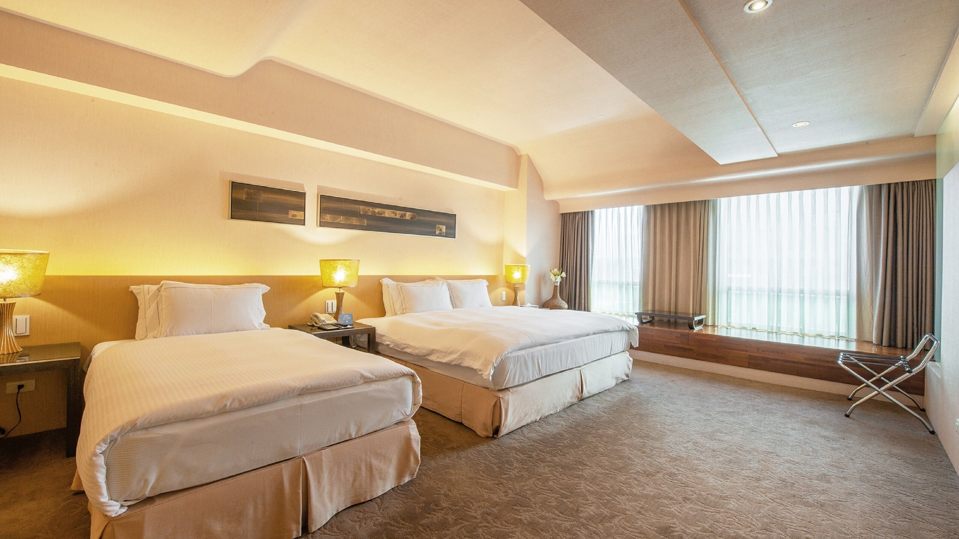 A hotel room with two beds, bedside lamps, a small table, a window with curtains, and a wall-mounted artwork.