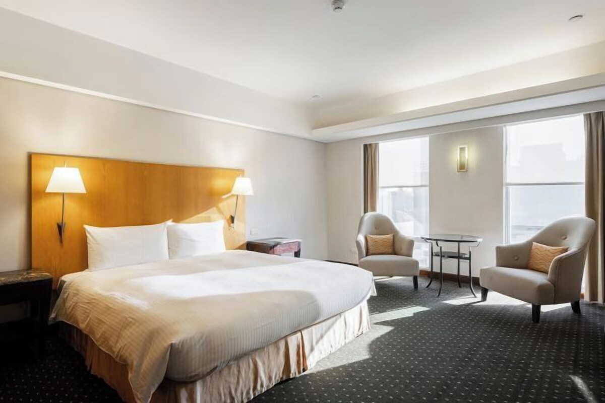 A hotel room with a large bed, two bedside lamps, a small table, two armchairs, and a view of the city through large windows.