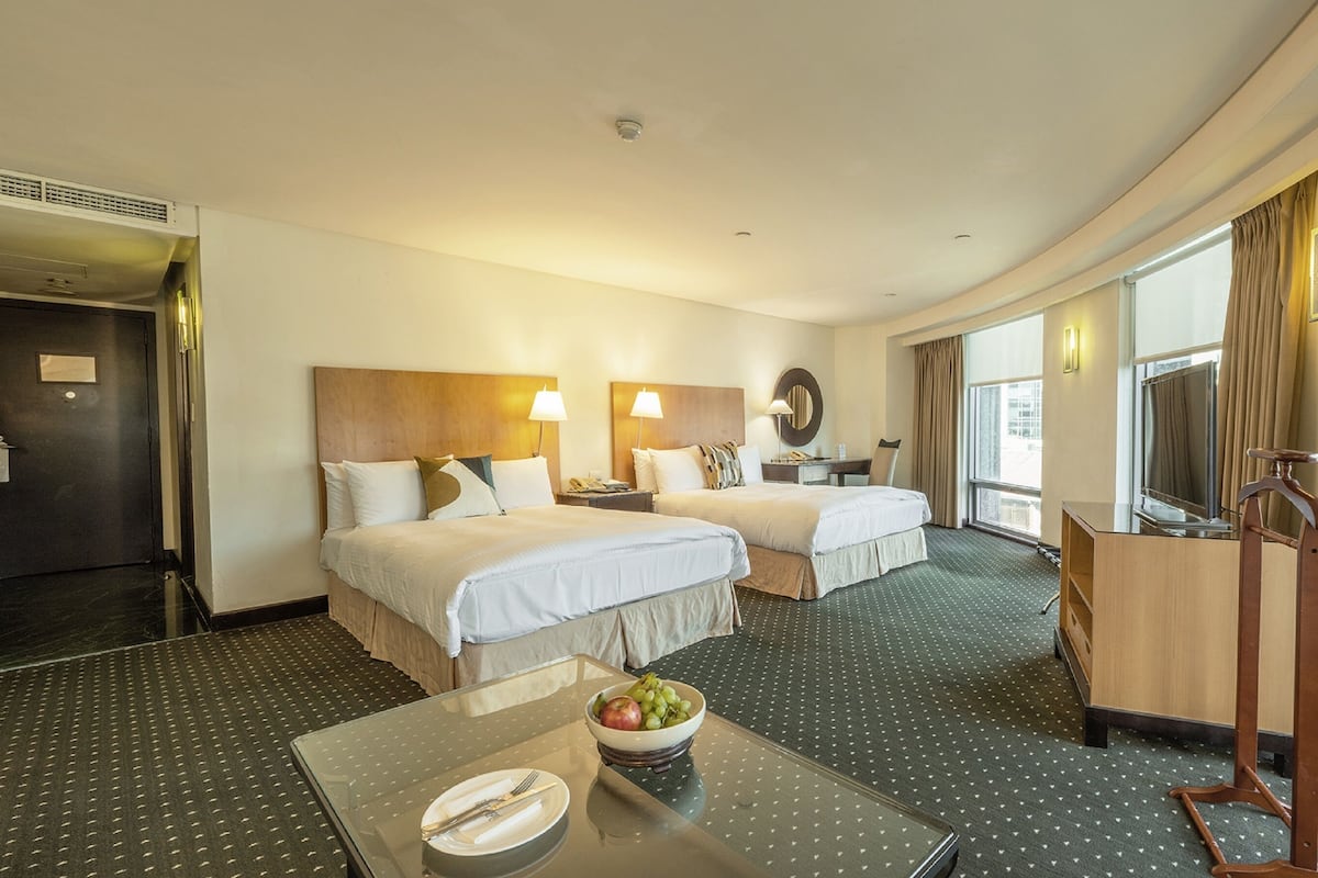 Quarto de hotel com duas camas, uma mesinha com uma cesta de frutas, uma escrivaninha com uma luminária e uma janela grande com cortinas.