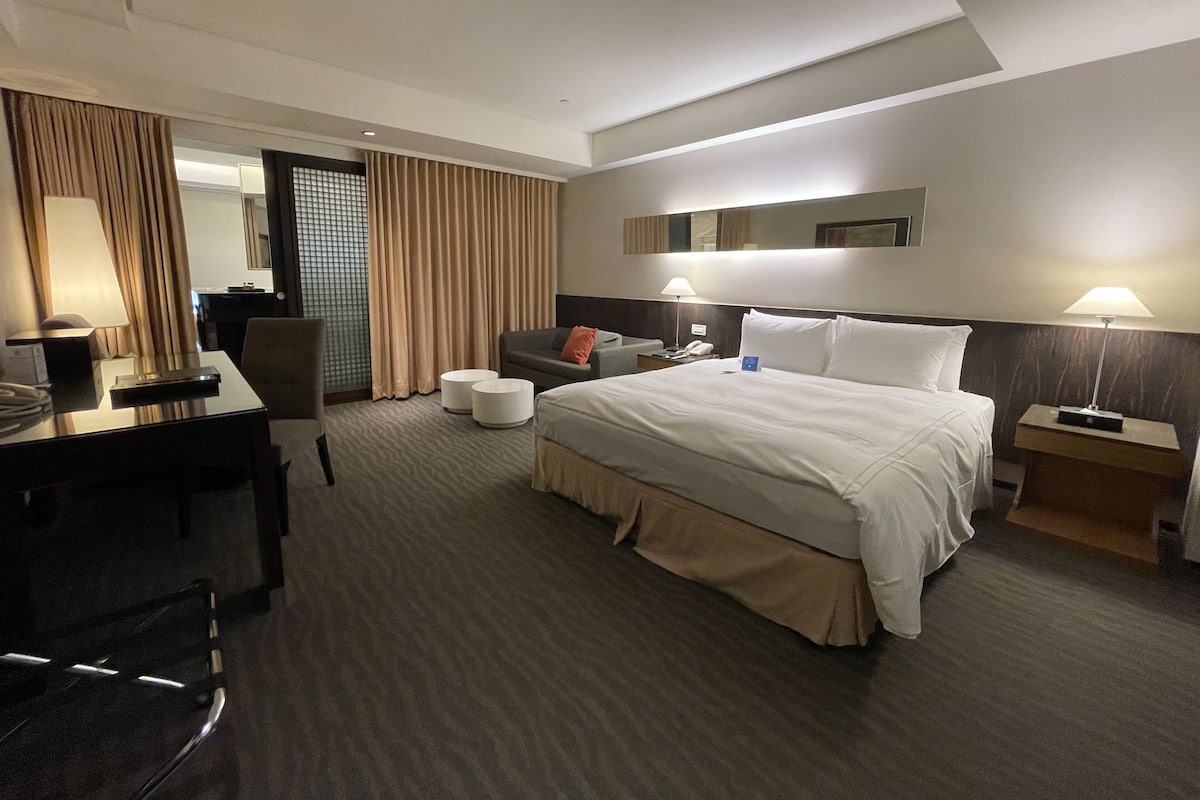Quarto de hotel moderno com uma cama grande, uma escrivaninha, uma cadeira, um sofá e uma mesinha de centro. Há duas luminárias de cabeceira e um espelho sobre a cama.