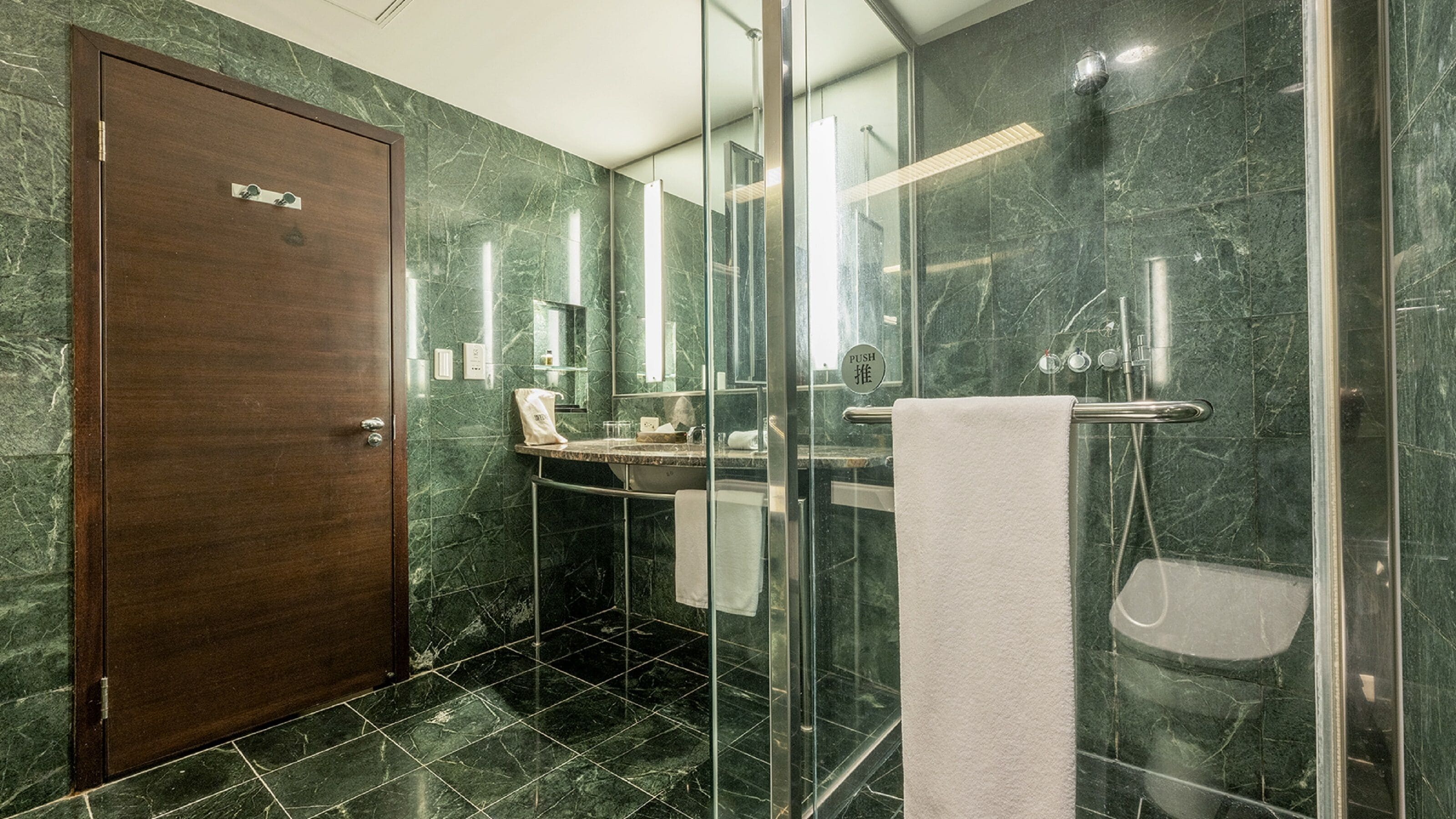 Un baño moderno con puerta de madera, cabina de ducha de vidrio, lavamanos con una toalla y un espejo.