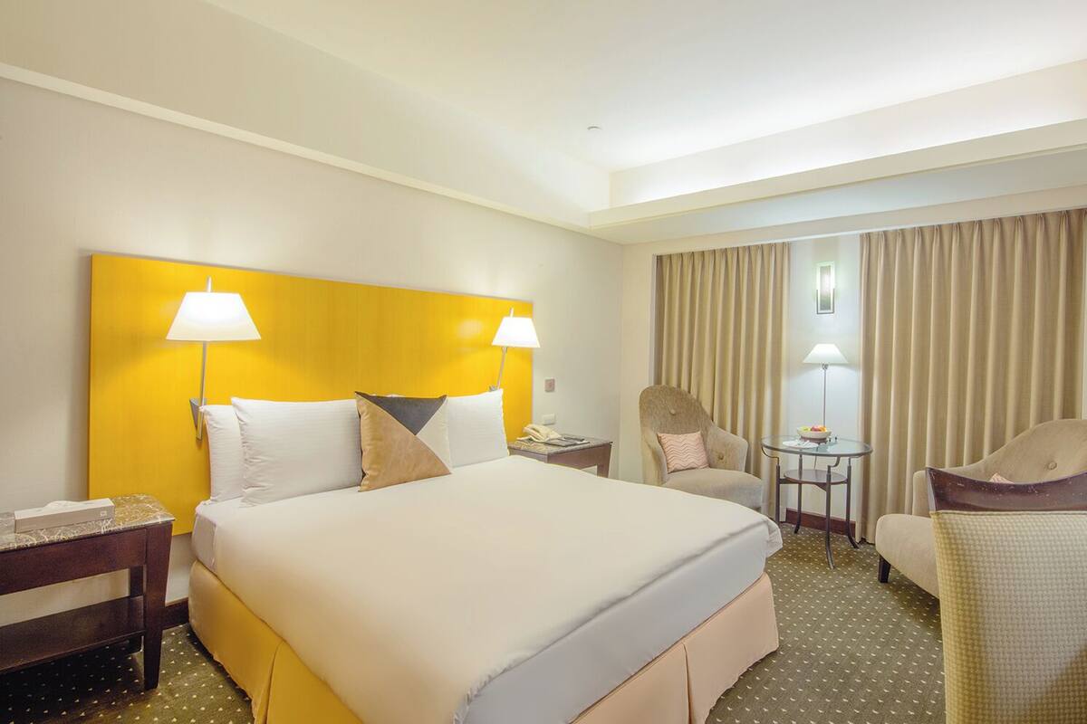 Quarto de hotel com uma cama grande, duas mesinhas de cabeceira, uma escrivaninha com cadeira, uma televisão e uma pequena mesa com abajur e uma tigela de frutas.