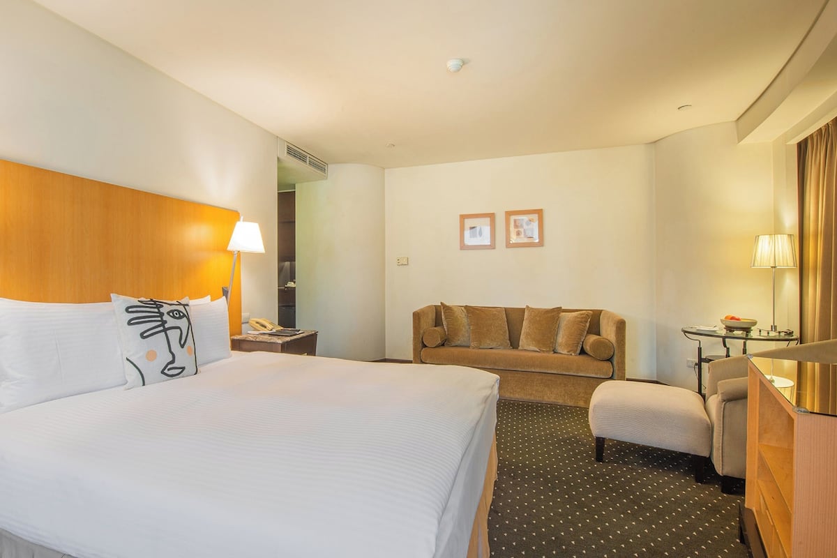Quarto de hotel com uma cama grande, duas luminárias de cabeceira, uma cabeceira de madeira, um sofá, uma poltrona, uma mesinha com uma cesta de frutas e um espelho.