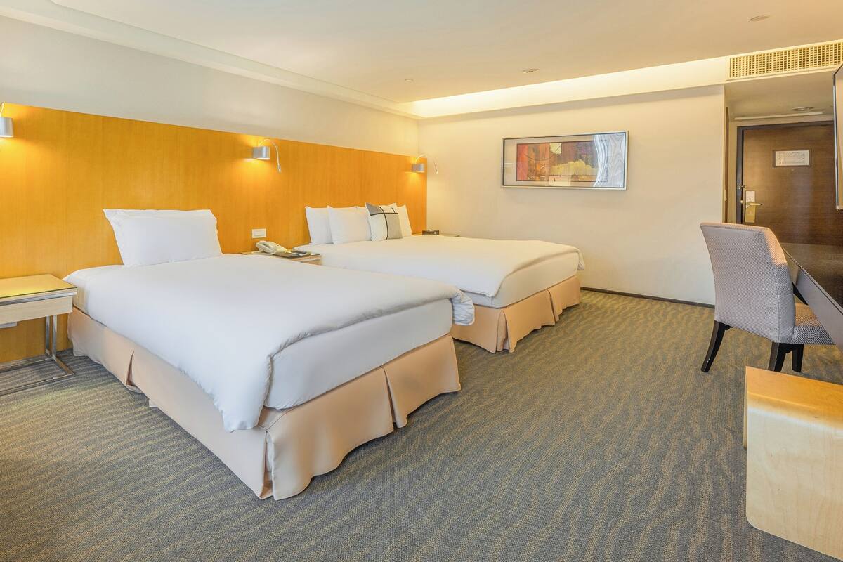 Quarto de hotel com duas camas, uma escrivaninha, uma cadeira e um quadro na parede.