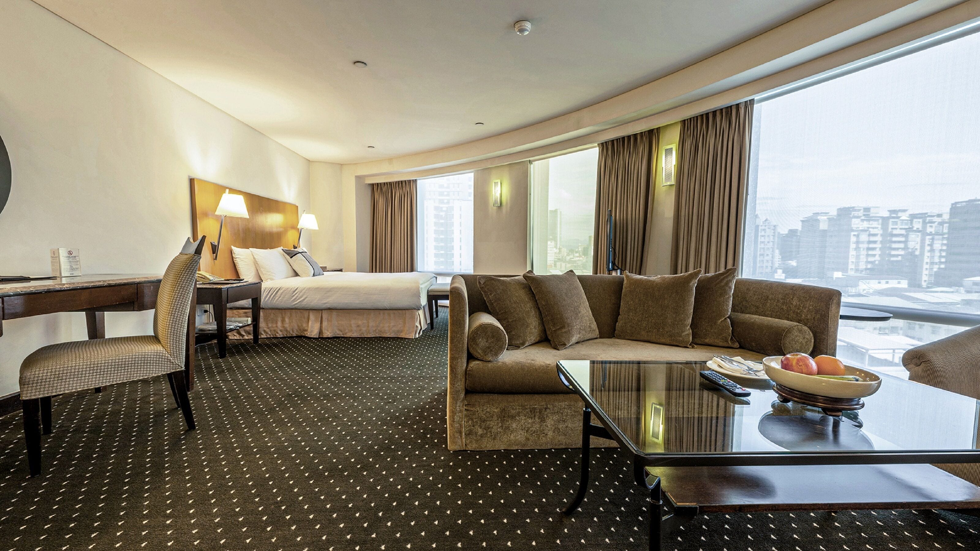 A hotel room with a bed, a sofa, a desk with a chair, a coffee table, and a view of the city.