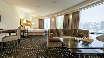A hotel room with a bed, a sofa, a desk with a chair, a coffee table, and a view of the city.