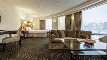A hotel room with a bed, a sofa, a desk with a chair, a coffee table, and a view of the city.