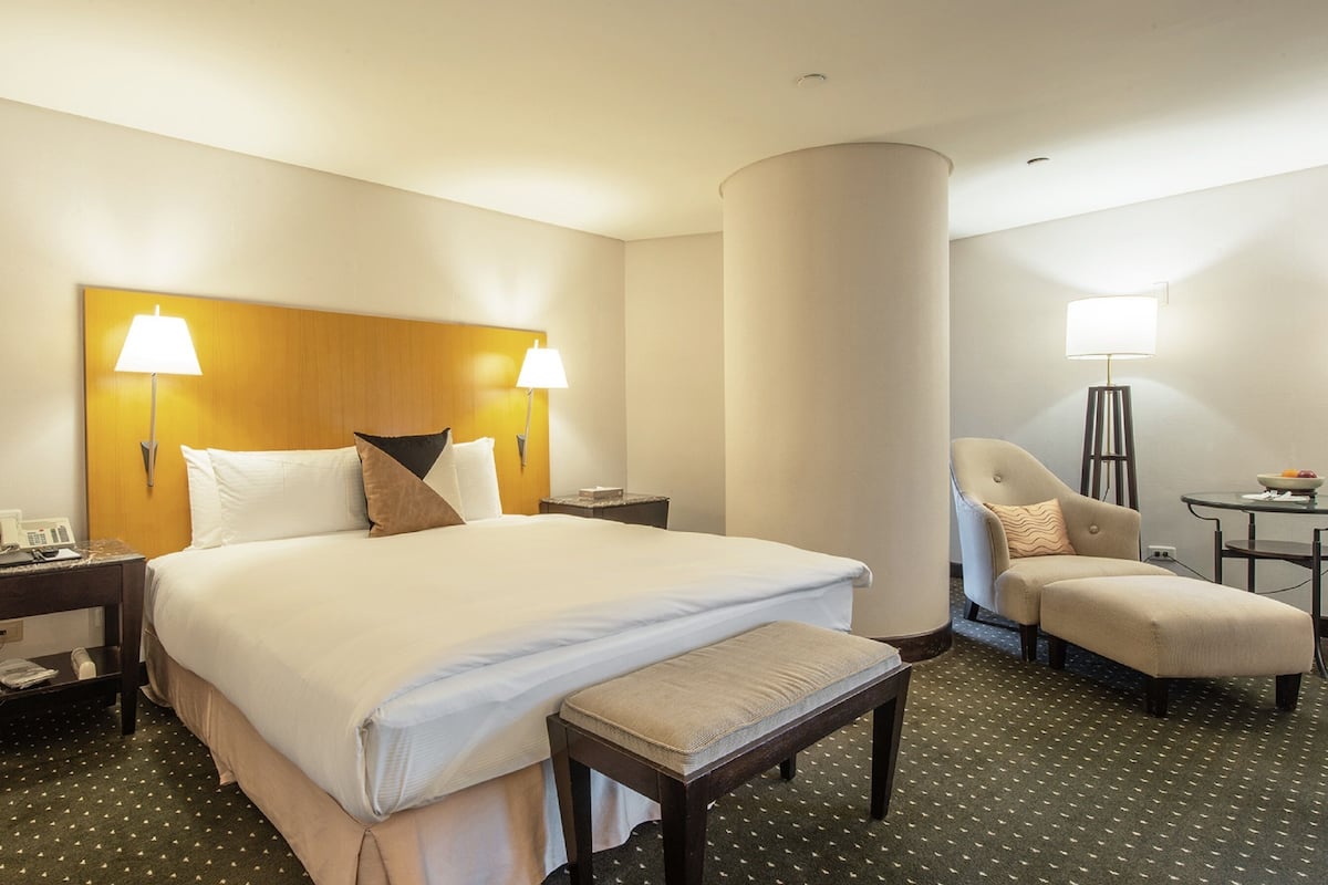 Quarto de hotel com uma cama grande, duas luminárias de cabeceira, uma cadeira, uma mesinha e um telefone no criado-mudo.