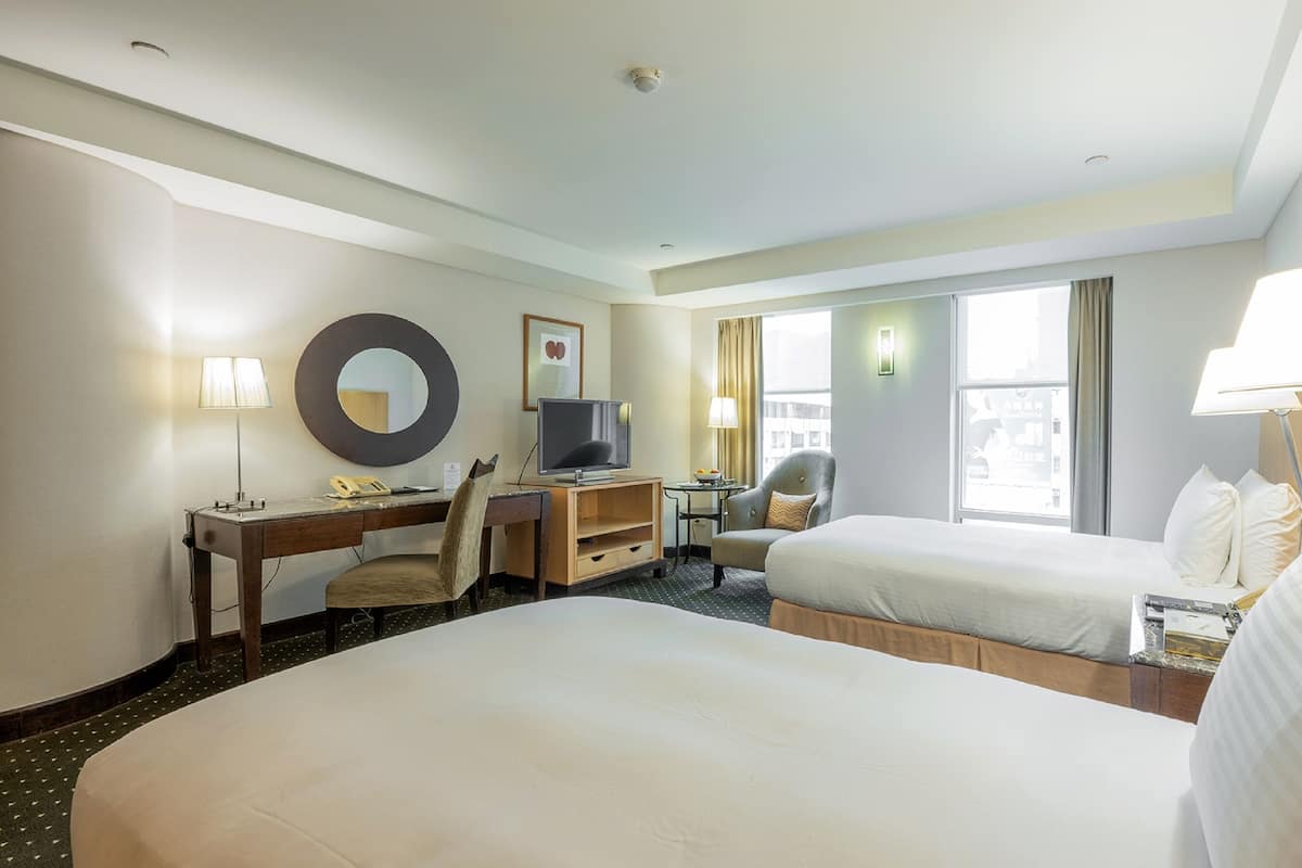 Quarto de hotel com duas camas, uma escrivaninha com computador, televisão e janela com cortinas.