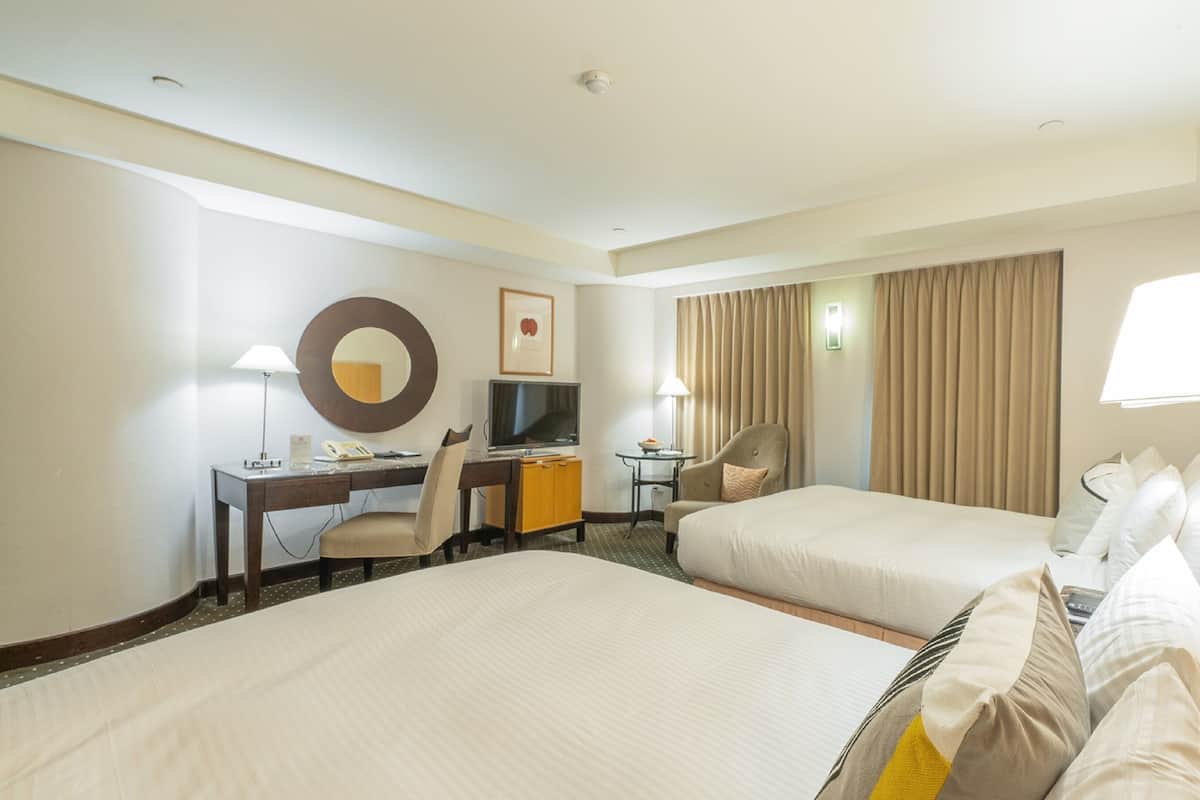 Quarto de hotel com duas camas, uma escrivaninha com cadeira, televisão, mesinha e candeeiro.