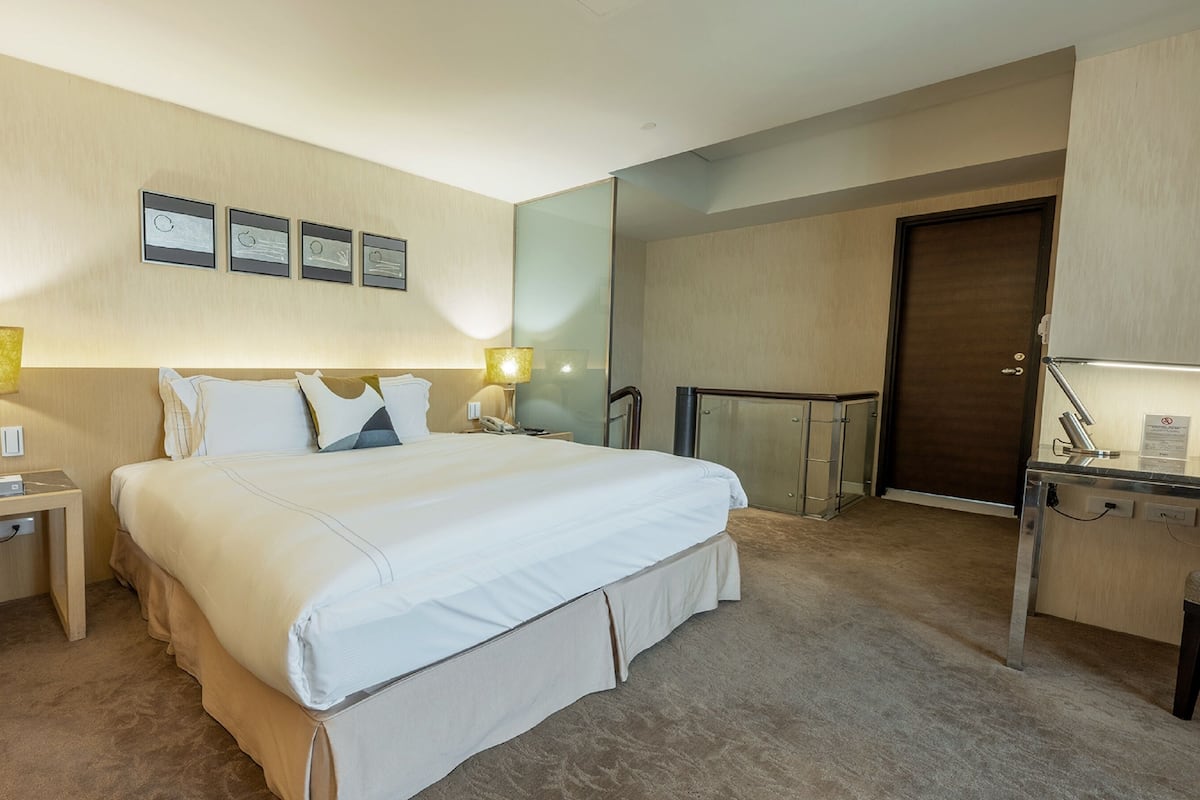 Quarto de hotel com uma cama grande, duas mesas de cabeceira com abajures, um espelho, um guarda-roupa e uma porta.