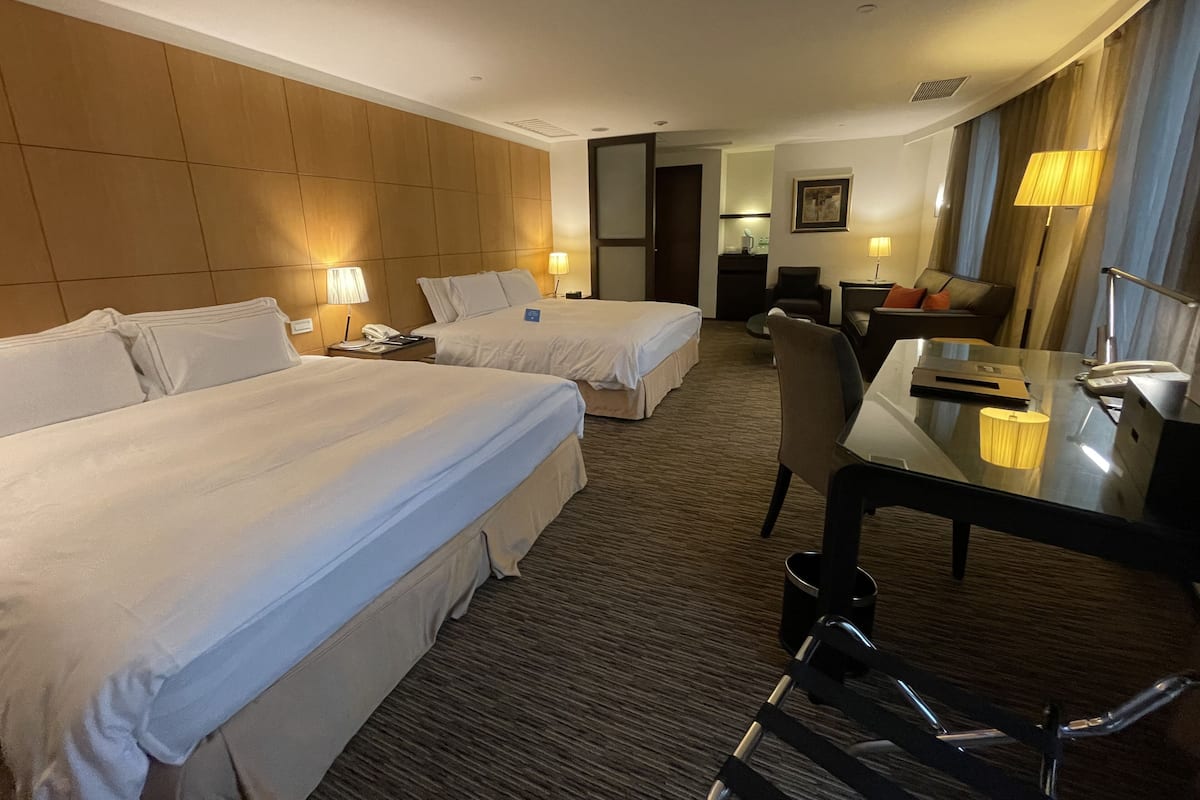 Quarto de hotel com duas camas, uma escrivaninha, uma cadeira e um pequeno espaço de estar com um sofá.