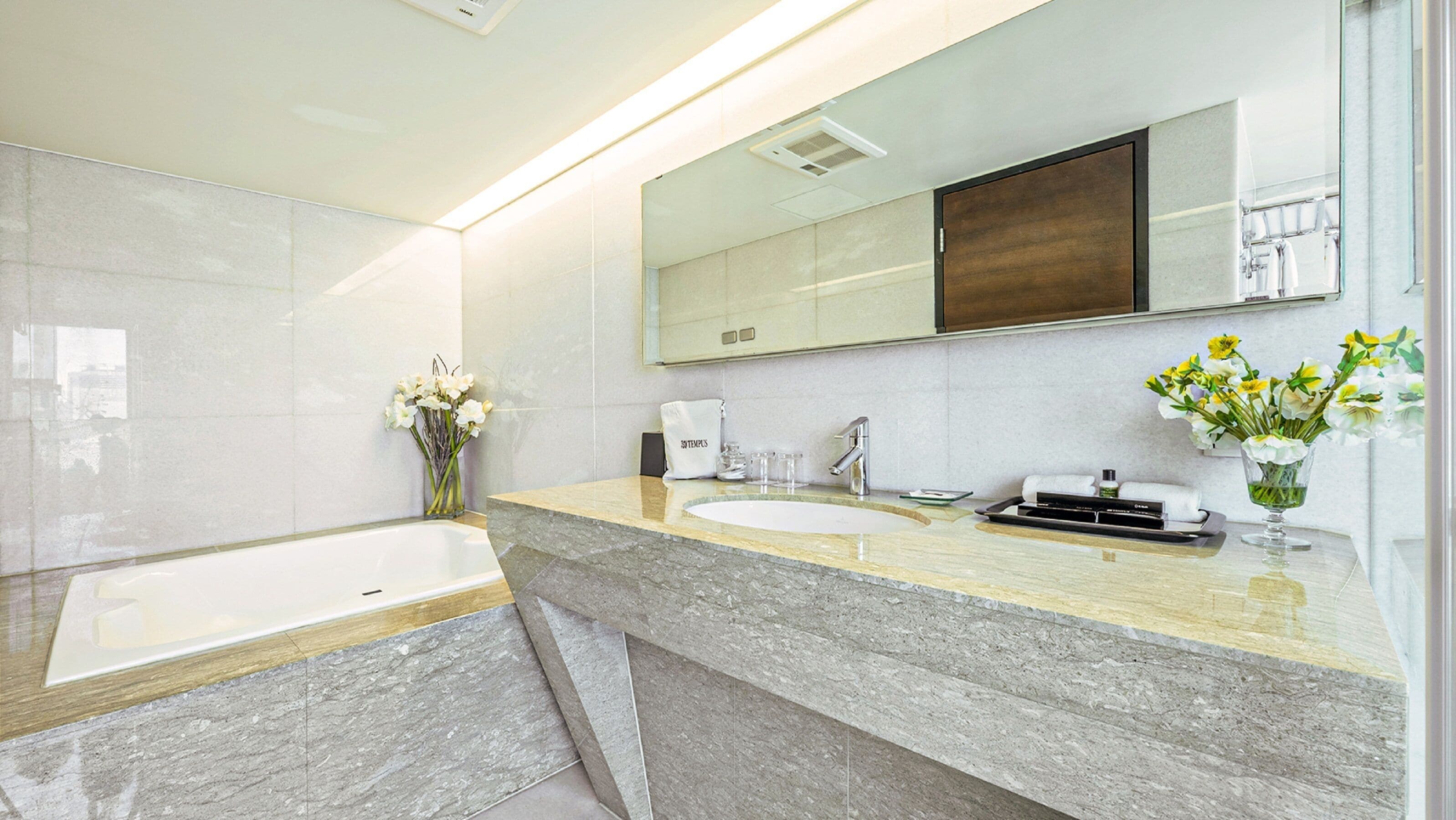 A modern bathroom with a bathtub, two sinks, a large mirror, and a vase with flowers.