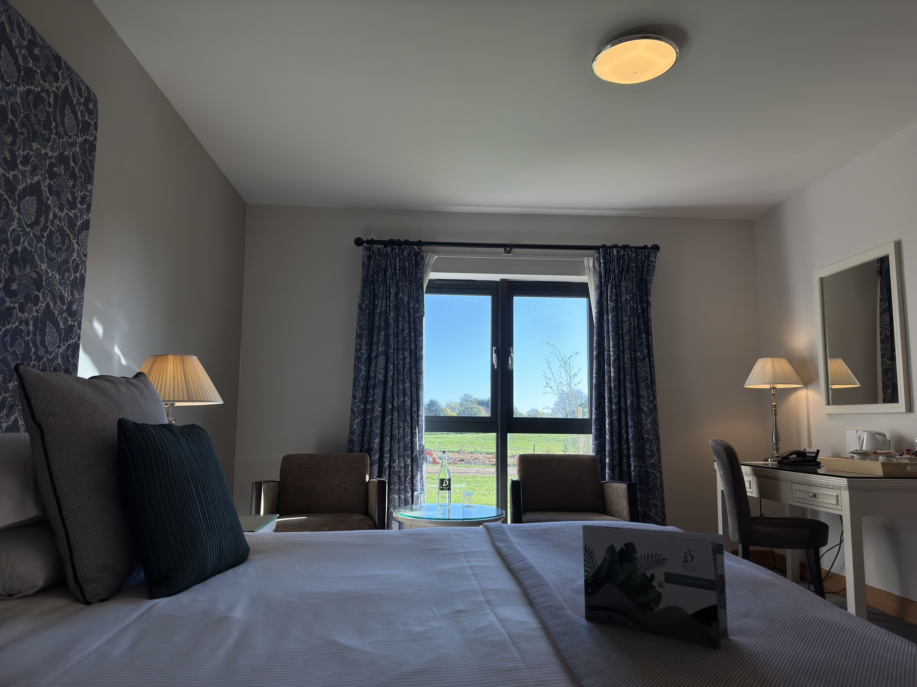 A hotel room with a bed, two chairs, a desk, a lamp, and a large window with curtains.
