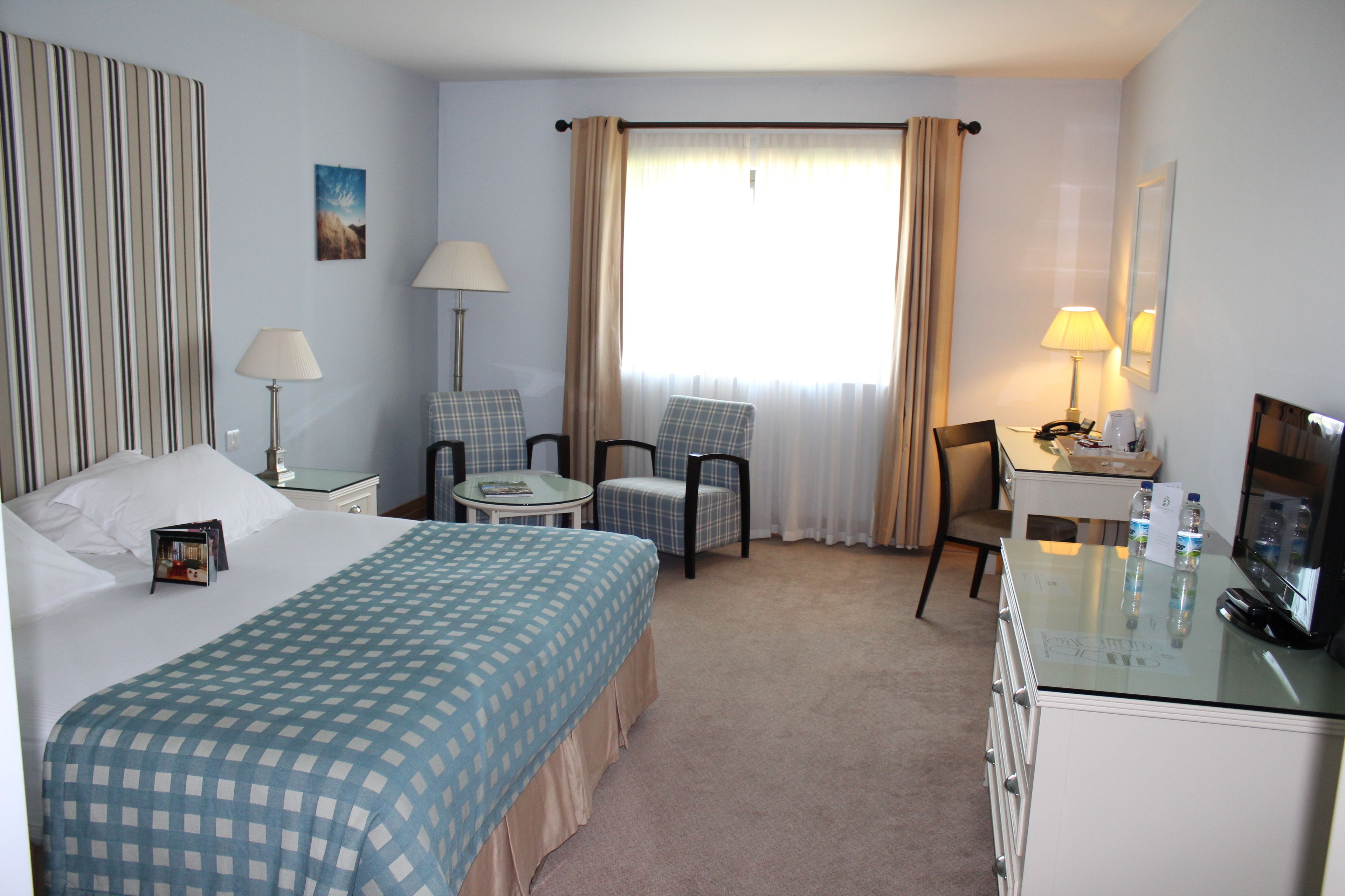 A hotel room with a bed, two chairs, a desk, a TV, and a window with curtains.
