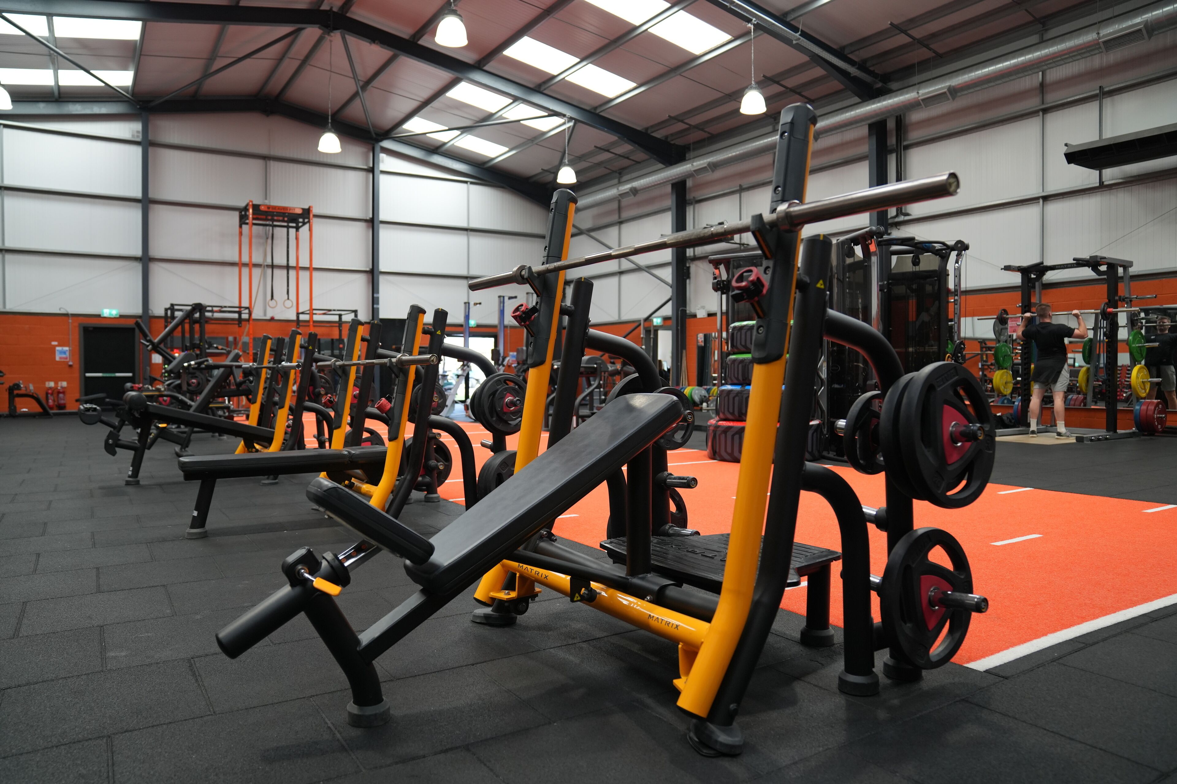 A gym with multiple weightlifting stations, including benches and squat racks, arranged in rows.