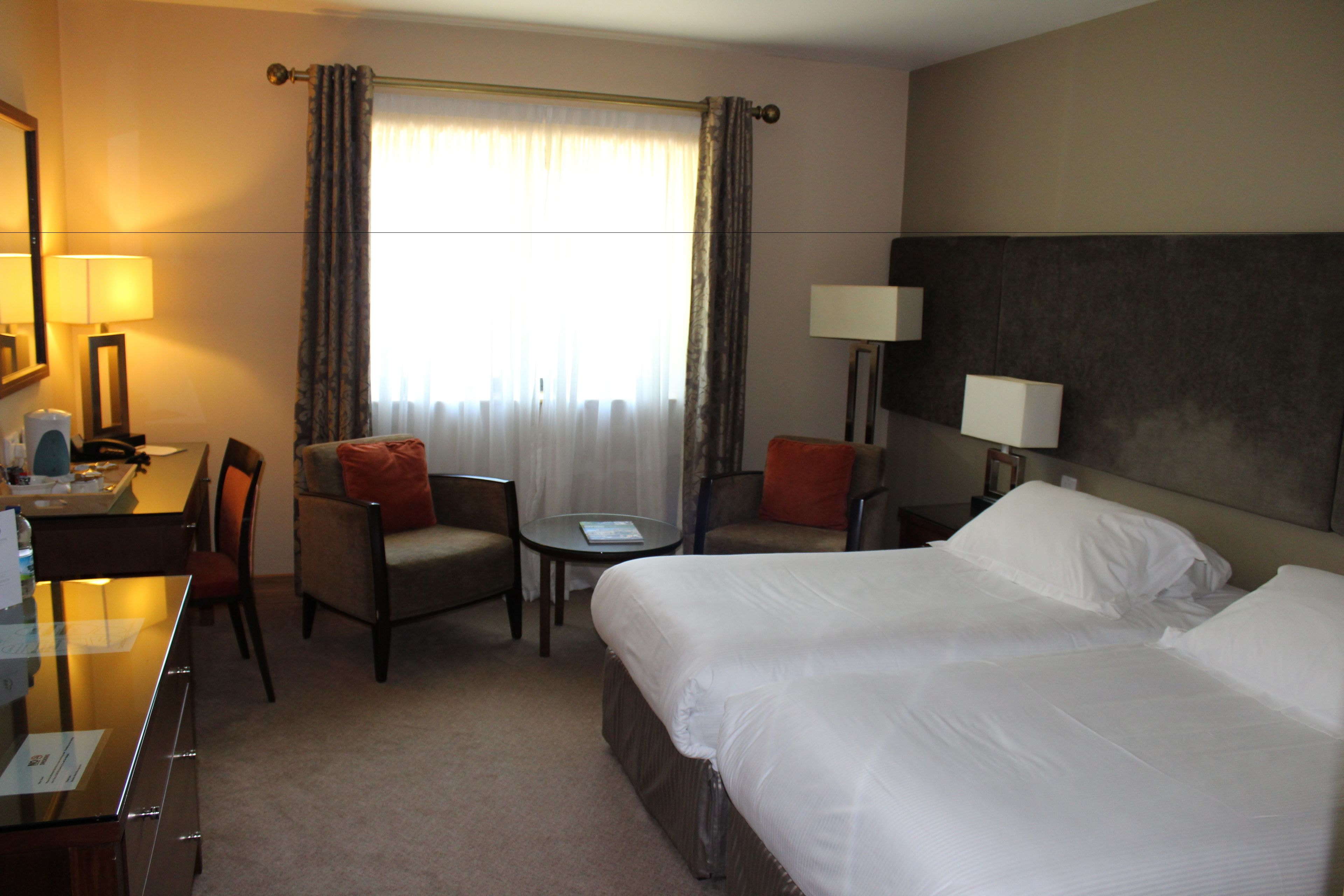 A hotel room with a large bed, a desk with a chair, a small table, two lamps, and a window with curtains.