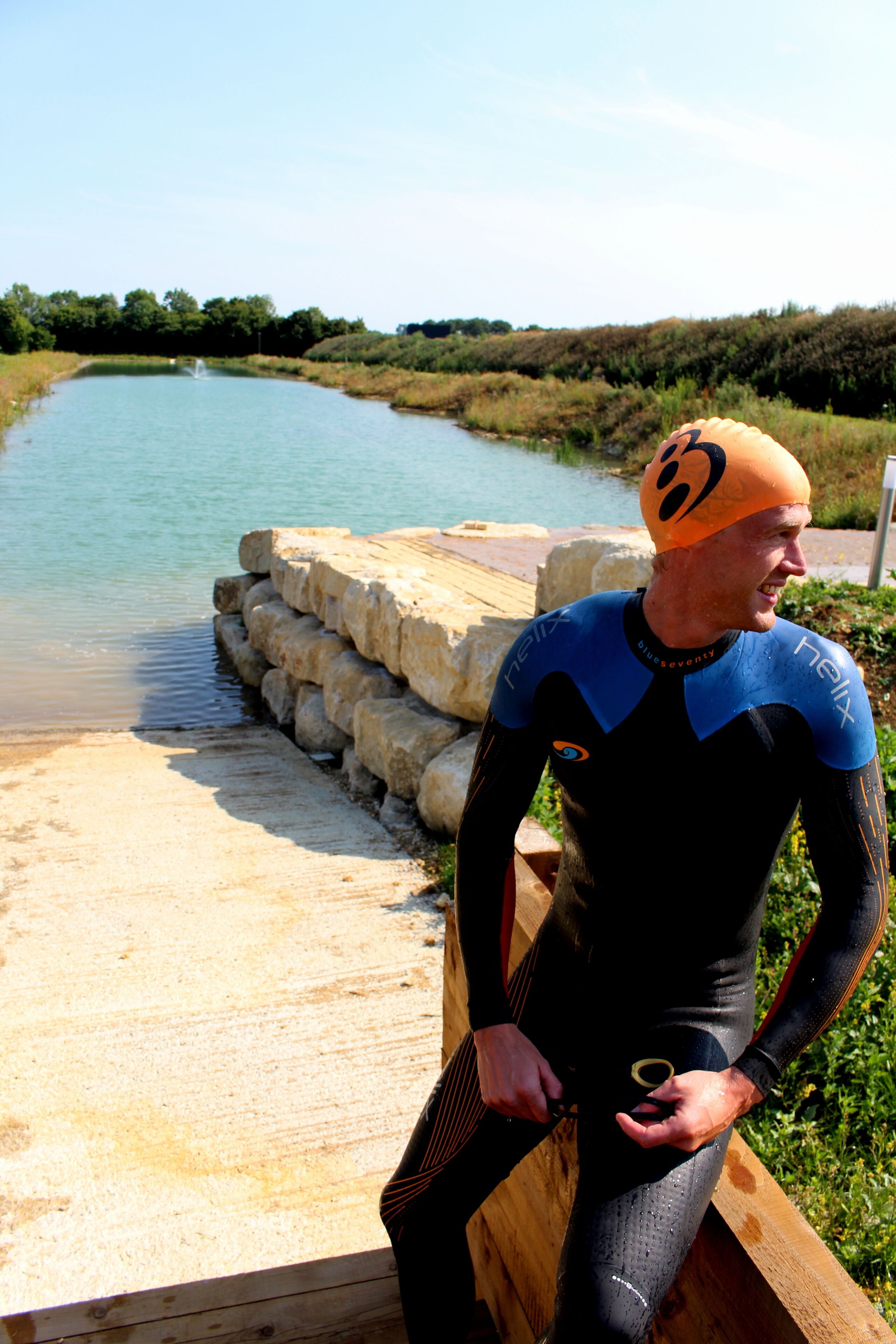 A person in a wetsuit sitting on a wooden dock by a calm body of water.