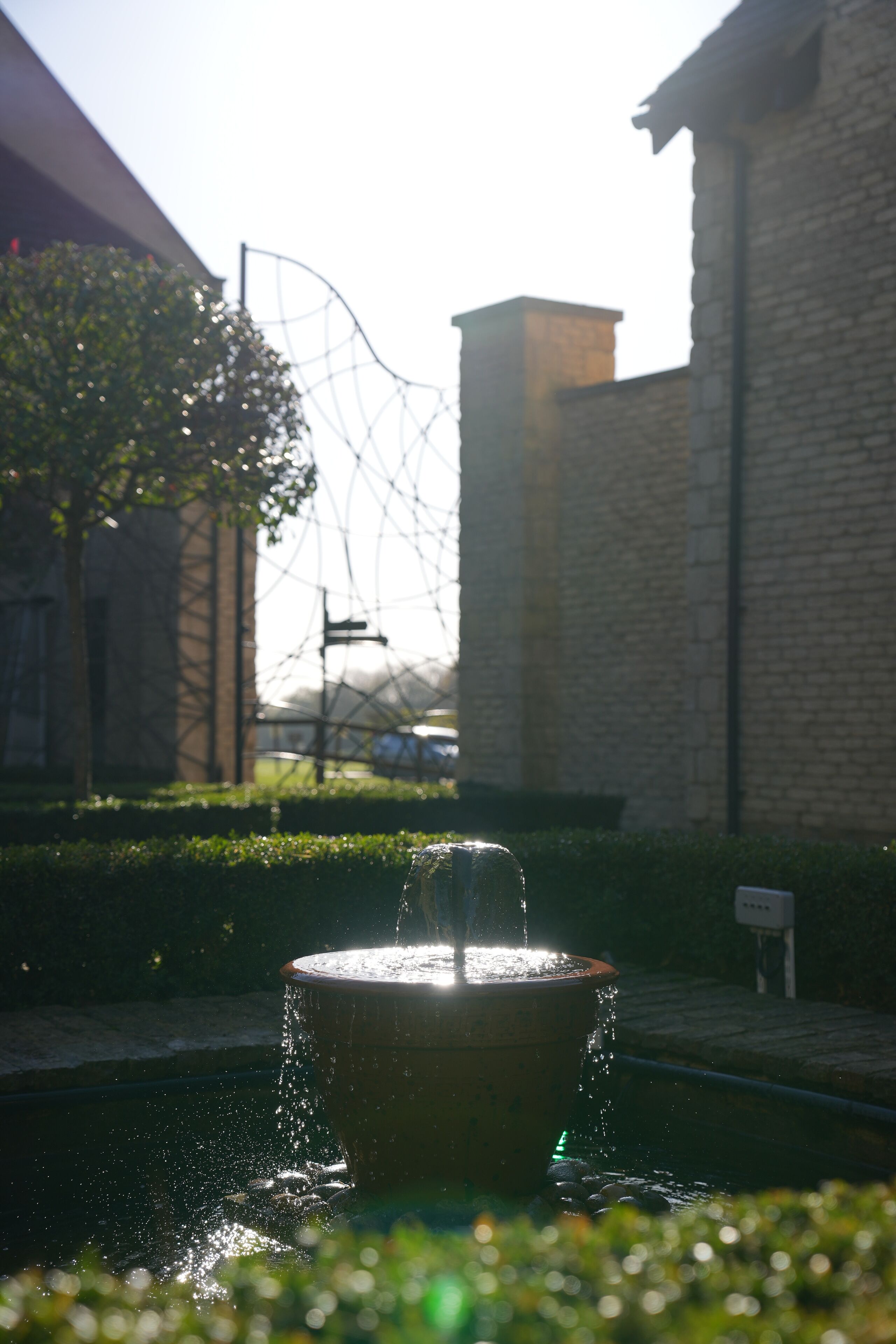 A fountain with water spouting from its center, surrounded by a paved area with a brick building in the background.