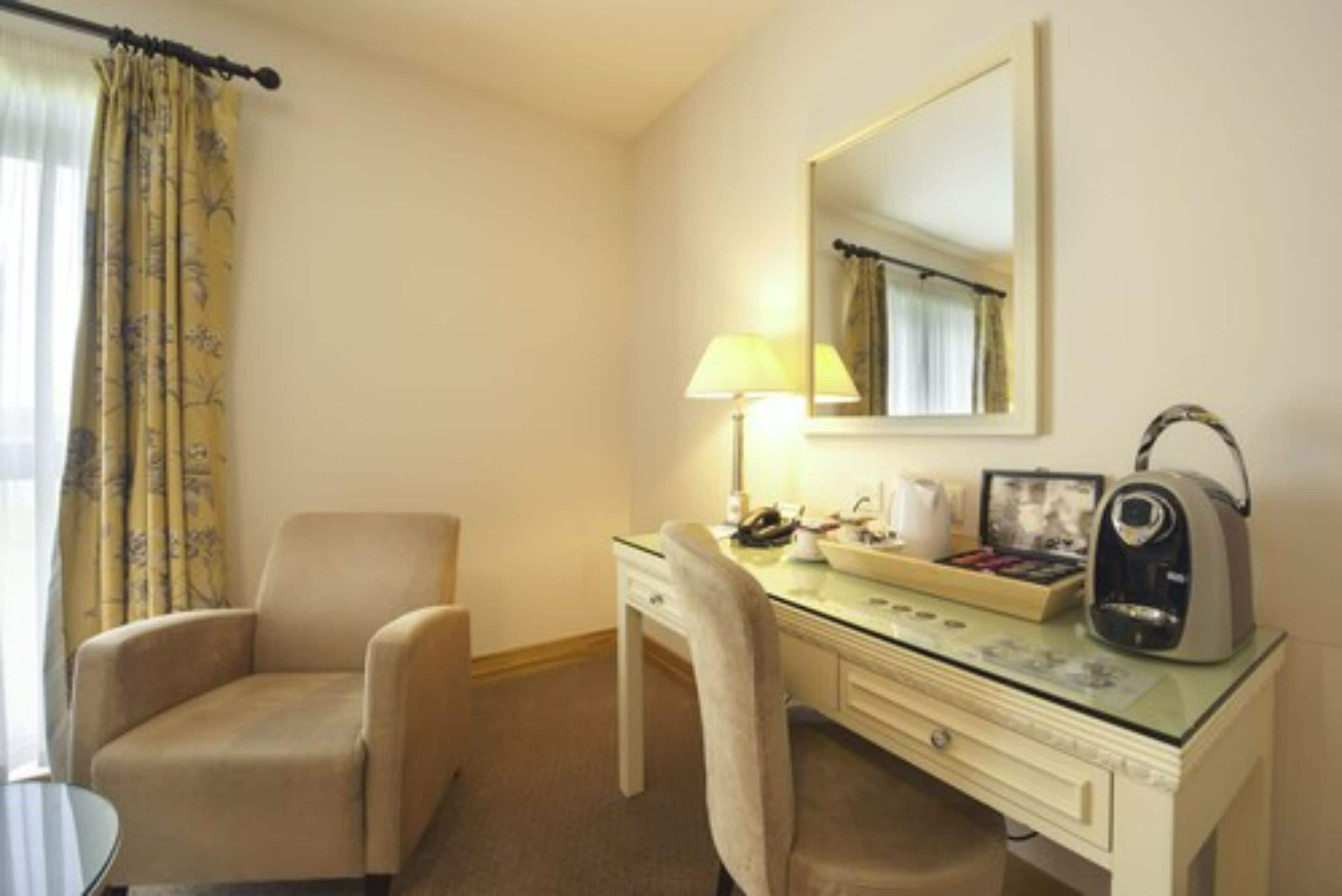 A hotel room with a beige armchair, a desk with a lamp, a coffee maker, and a mirror.