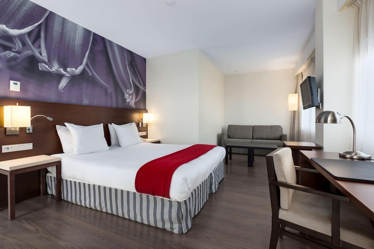 A hotel room with a large bed, a desk, a chair, a sofa, and a TV mounted on the wall.