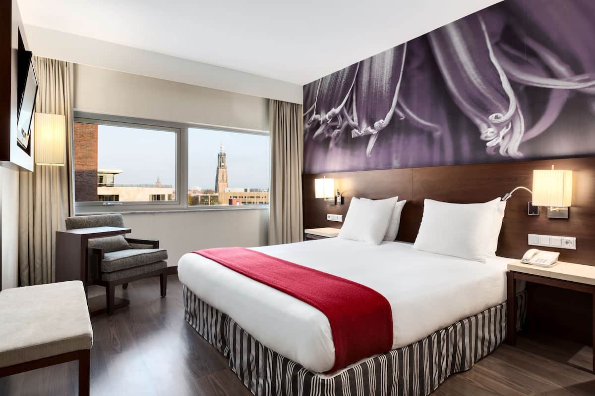 A hotel room with a large bed, a desk, a chair, and a view of a cityscape.