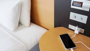 A hotel room with a bed, a bedside table with a charging phone, and a digital thermostat.