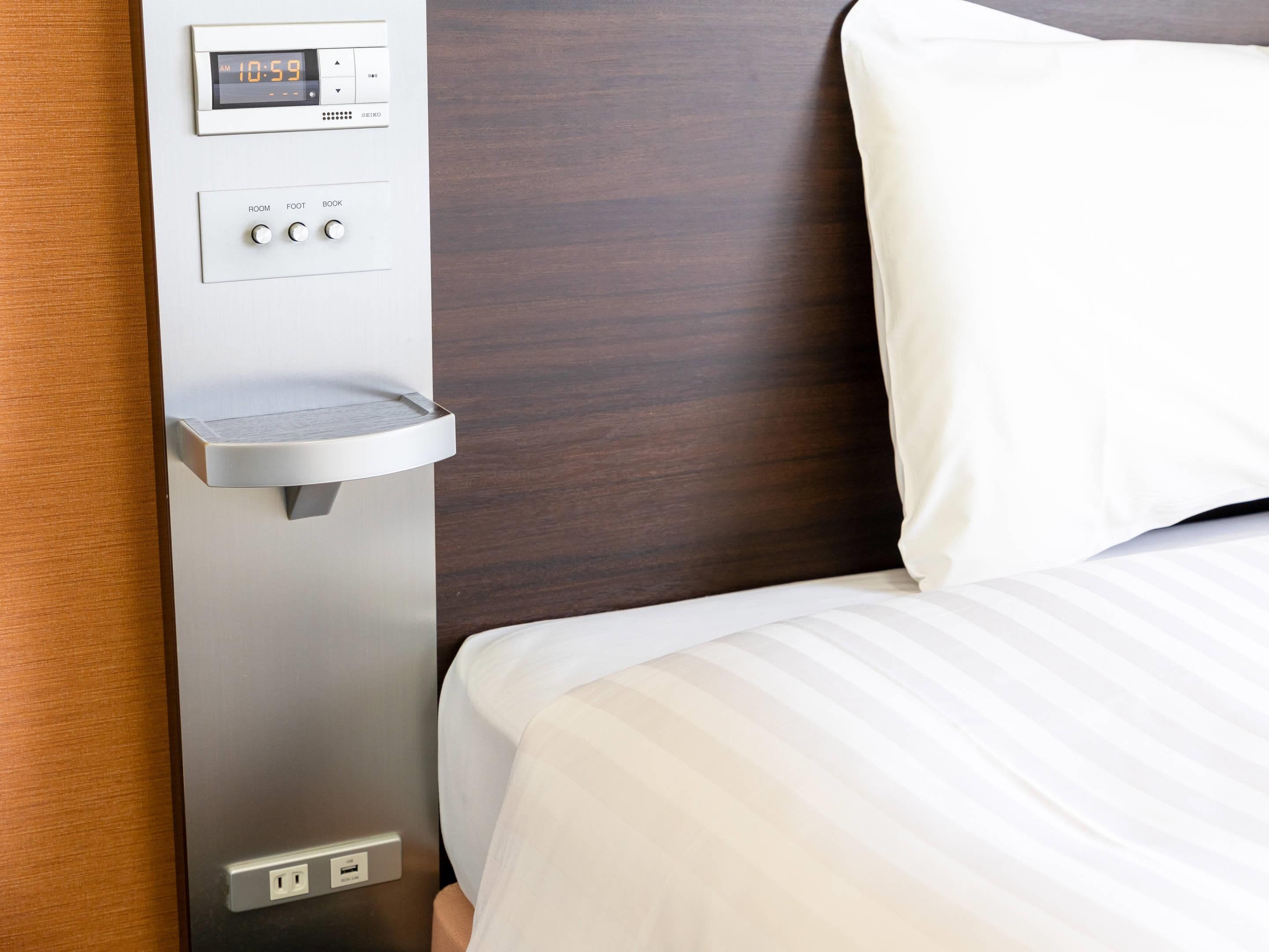 A hotel room with a bed, a bedside table with a digital clock, and a power outlet.