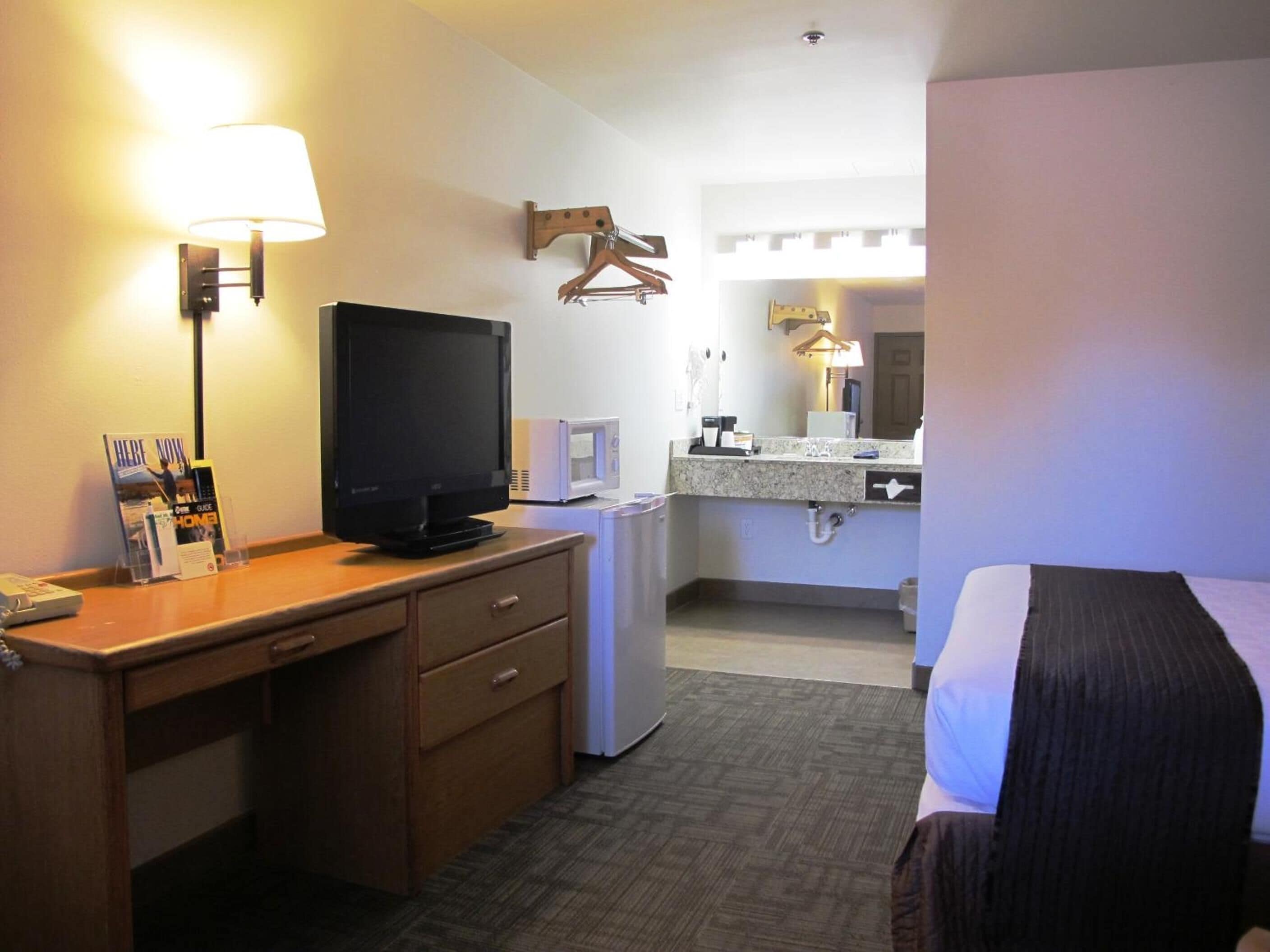 A hotel room with a bed, a desk with a television, a microwave, a small refrigerator, a mirror, and a hanging coat rack.